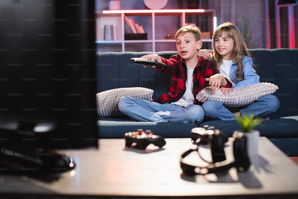 Happy caucasian kids using remote control for choosing cartoon on TV while sitting on comfy couch. Brother and sister relaxing and playing together at home.
