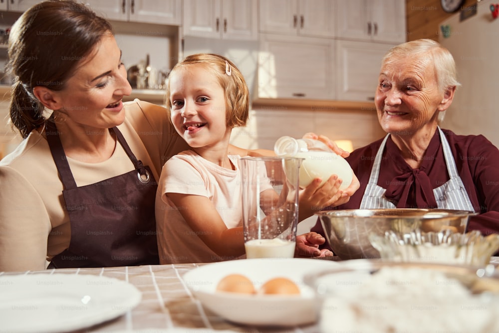 Hija vertiendo leche en un gran vaso medidor y mostrando la lengua a la cámara mientras la madre y la abuela la miran