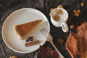 Pumpkin pie slice on modern plate and hot coffee on rustic table with linen napkin, autumn flowers and leaves, anise and cinnamon. Homemade pumpkin tart recipe. Top view. Happy Thanksgiving