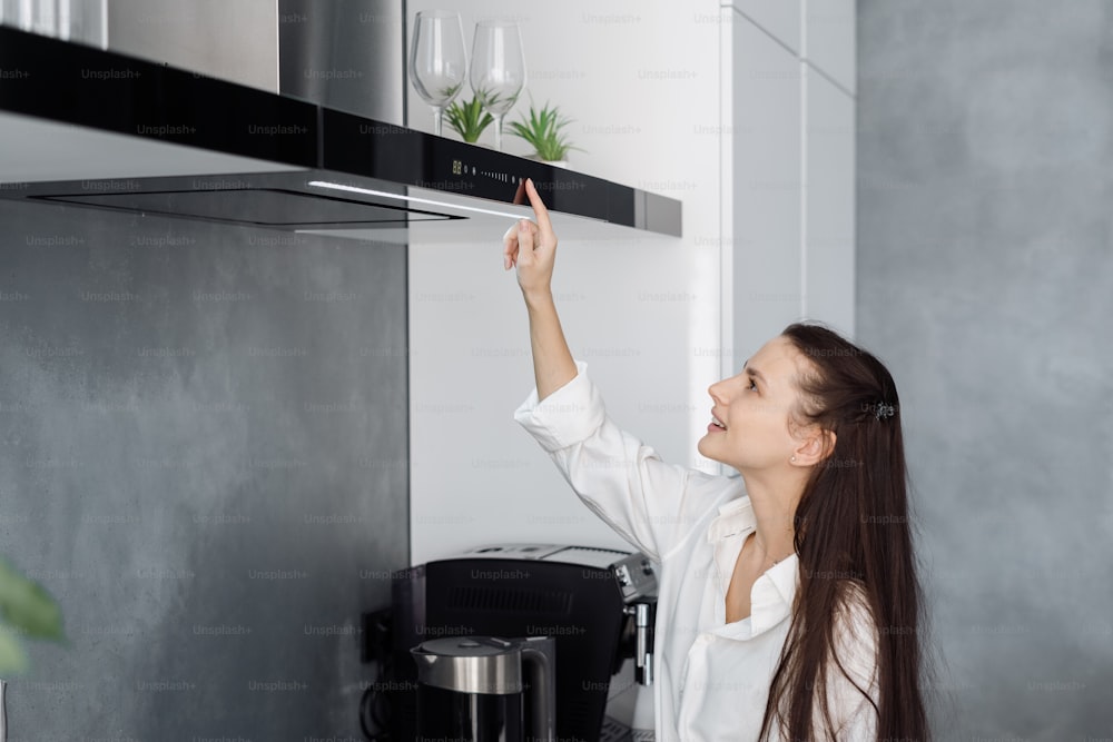 Jovem mulher feliz dona de casa usando exaustão de cozinha preta com tela sensível ao toque no painel de controle acima do fogão elétrico enquanto prepara na cozinha minimalista totalmente mobiliada com eletrodomésticos integrados