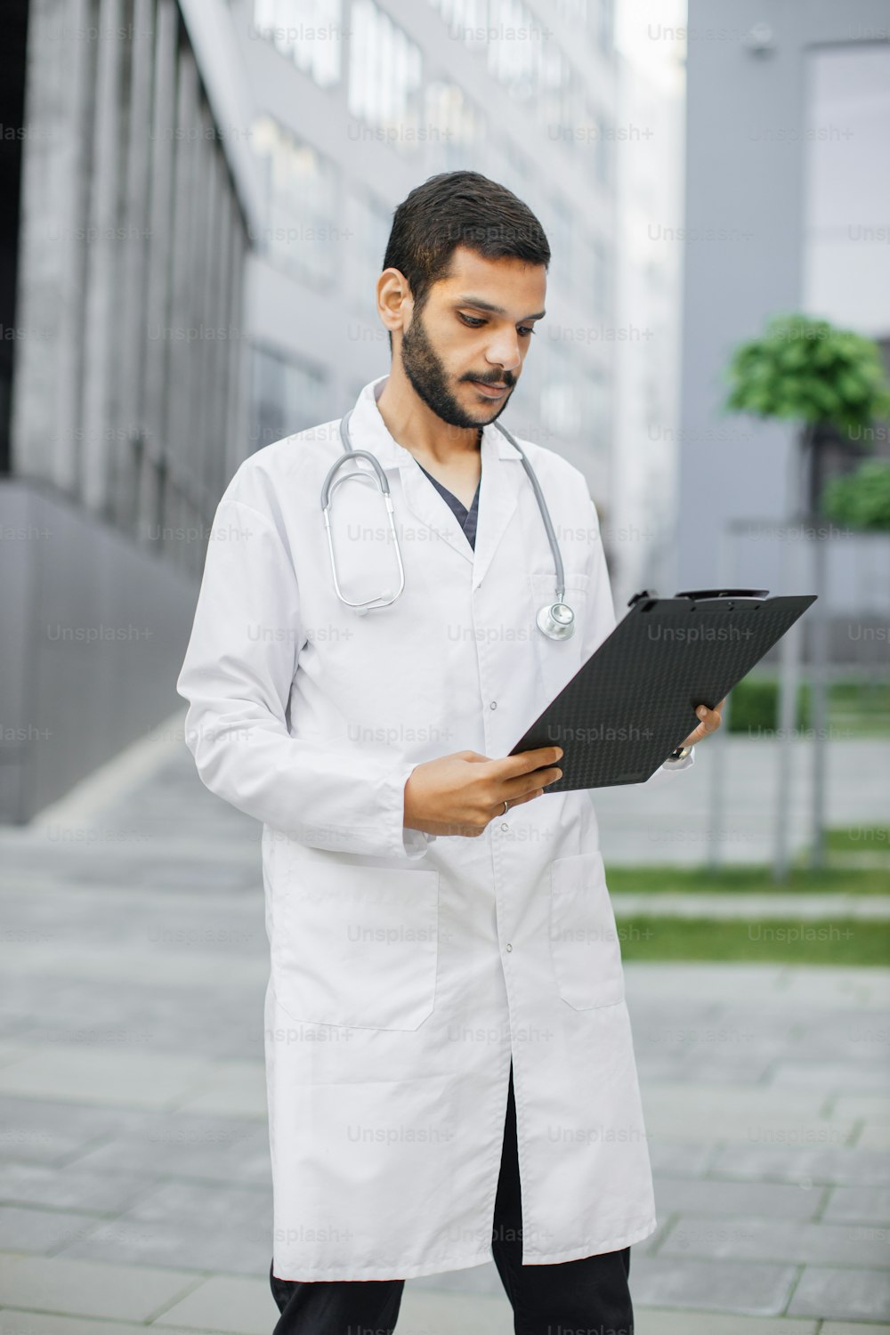 Portrait of young smart focused male medical intern doctor reading the data on folder clipboard, standing outside modern hospital building. Medicine, education and healthcare concept
