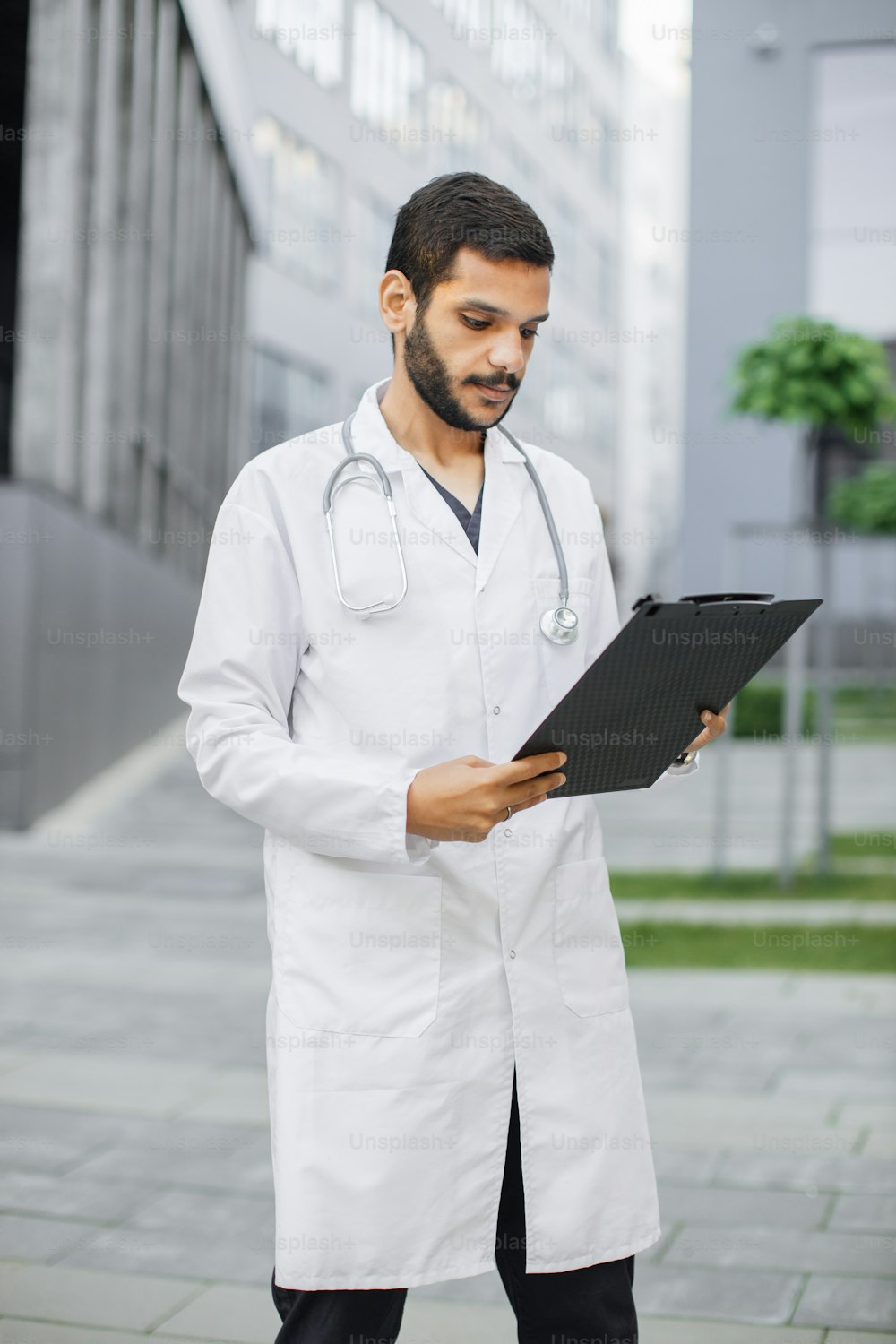 Porträt eines jungen, intelligenten, fokussierten männlichen medizinischen Praktikanten, der die Daten auf dem Ordner-Klemmbrett liest und vor dem modernen Krankenhausgebäude steht. Medizin-, Bildungs- und Gesundheitskonzept