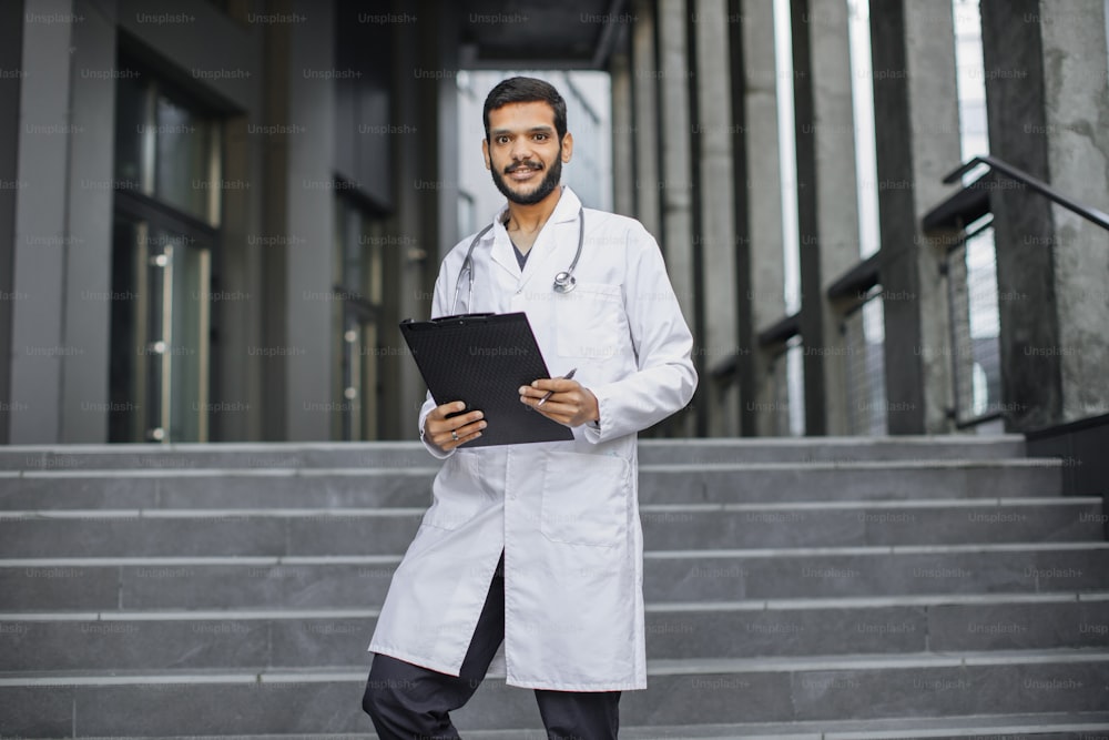 Close up front view of pleasant young Middle Eastern ethnic doctor in white gown, holding a folder, standing on stairs of modern clinic outdoors, smiling and looking at camera