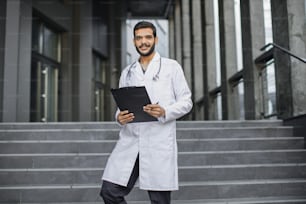 Vue rapprochée de face d’un jeune médecin ethnique du Moyen-Orient agréable en robe blanche, tenant un dossier, debout sur les escaliers d’une clinique moderne à l’extérieur, souriant et regardant la caméra