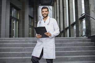 Vista frontale ravvicinata di piacevole giovane medico etnico mediorientale in camice bianco, in possesso di una cartella, in piedi sulle scale della clinica moderna all'aperto, sorridente e guardando la macchina fotografica