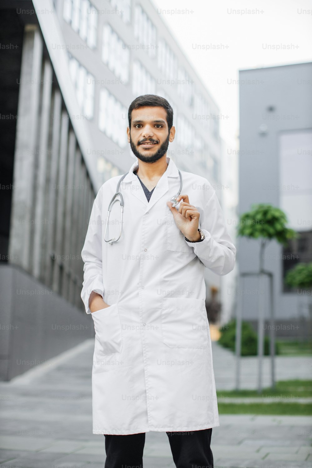 Close up outdoor shot of smiling friendly young Arabian Indian male doctor or student in white medical coat, standing outside modern clinic or campus with stethoscope around neck