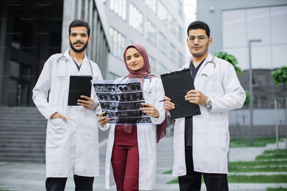 Vista frontal de três médicos, uma senhora muçulmana de hijab e dois homens árabes, trabalhando em frente à clínica. Mulher com tomografia computadorizada, homens com tablet pc e prancheta, discutindo o diagnóstico do paciente