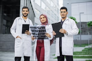 Vue de face de trois médecins, d’une femme musulmane en hijab et de deux hommes arabes, travaillant devant la clinique. Femme avec tomodensitométrie, hommes avec une tablette et un presse-papiers, discutant du diagnostic du patient