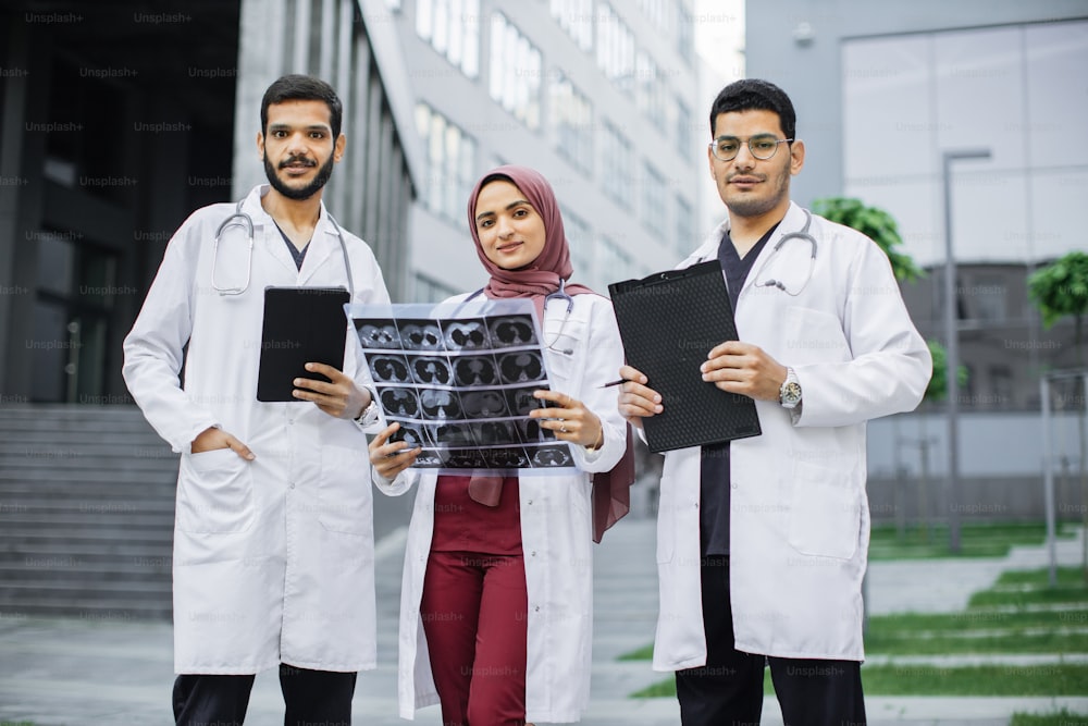 Front view of three doctors, Muslim lady in hijab and two Arabian men, working in front of the clinic. Woman with CT scan, men with tablet pc and clipboard, discussing patient's diagnosis