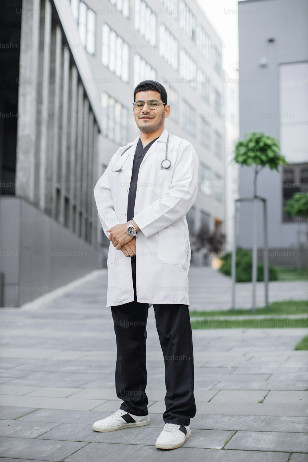 Um jovem e bonito médico da Arábia Saudita sorrindo do lado de fora do hospital. Retrato de corpo inteiro do estudante de medicina do sexo masculino em jaleco branco, posando na frente do edifício moderno da universidade ou clínica