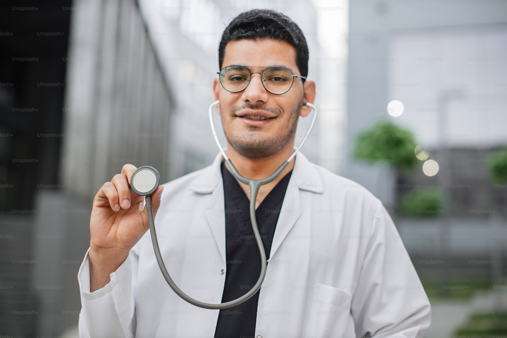 Portrait of smiling friendly male Arab hindu doctor or medical student standing outside modern hospital, demonstrating his stethoscope to camera. Selective focus on stethoscope