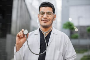 미소 짓는 친절한 남성 아랍 힌두교 의사 또는 의대생이 현대 병원 밖에 서서 청진기를 카메라에 시연하는 초상화. 청진기에 대한 선택적 초점