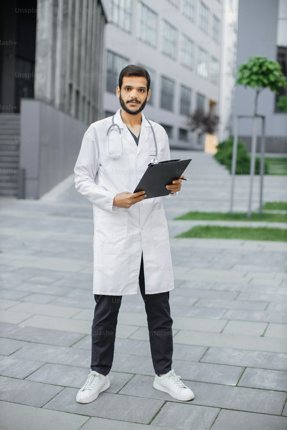 Full length portrait of young handsome bearded Arabian man doctor healthcare worker in white coat, standing with clipboard folder in hands outdoors in front of modern hospital or clinic