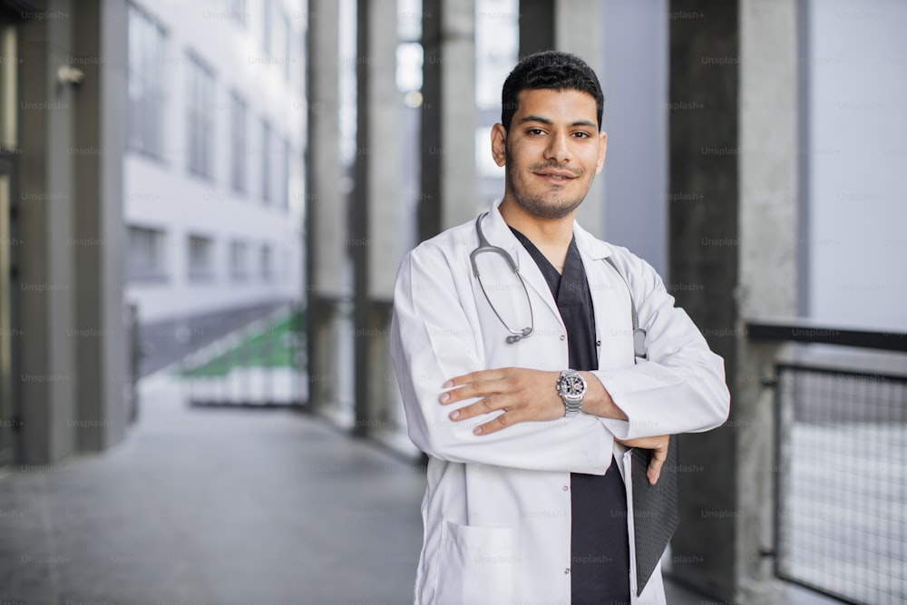 Horizontal waist-up portrait of a handsome male Arab Malay doctor, wearing white coat, standing with arms crossed outdoors on the background of clinic building. Close up, copy space