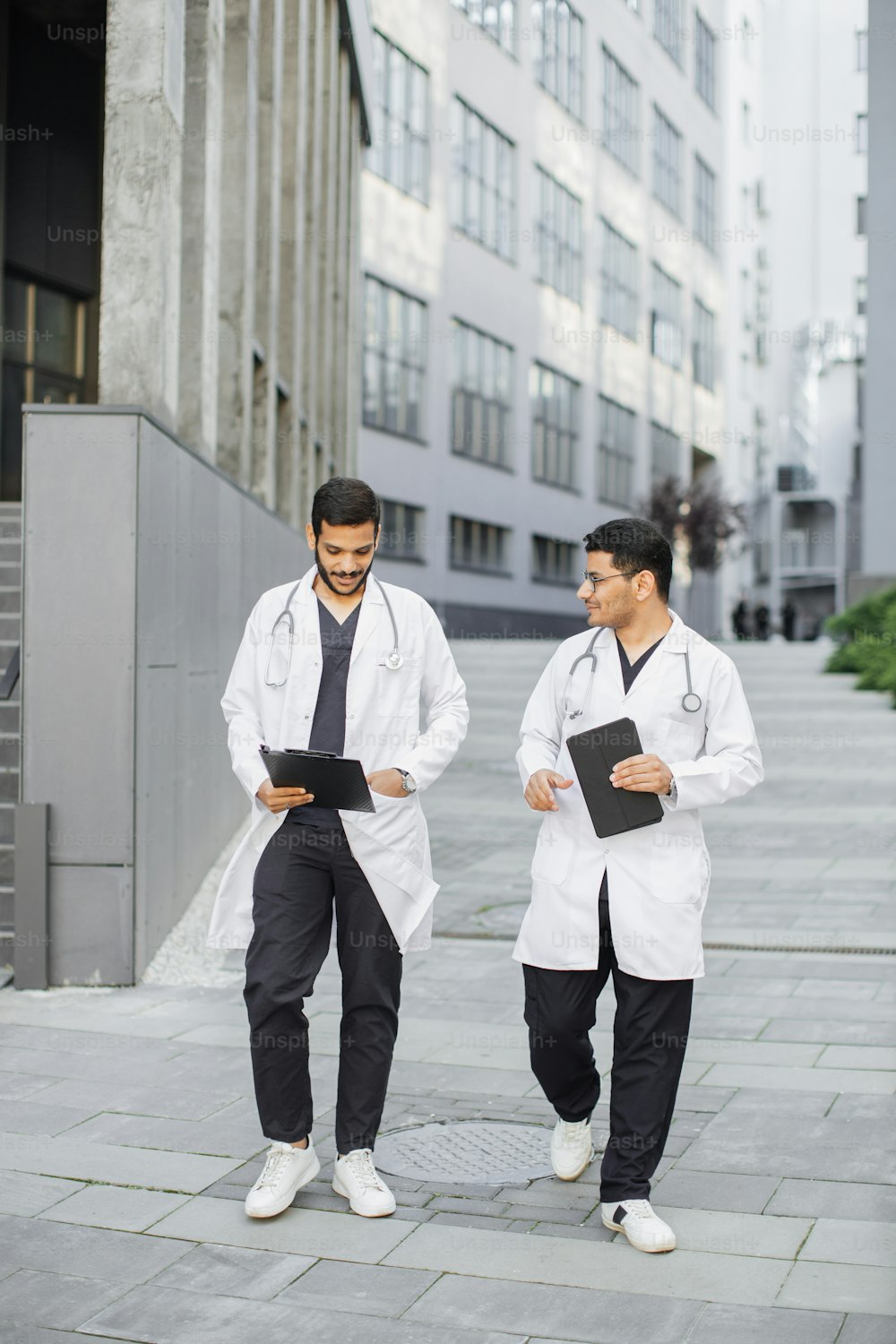 Two male Indian Arabian doctors in medical apparel, discussing patient's diagnosis, walking outside on the background of modern hospital