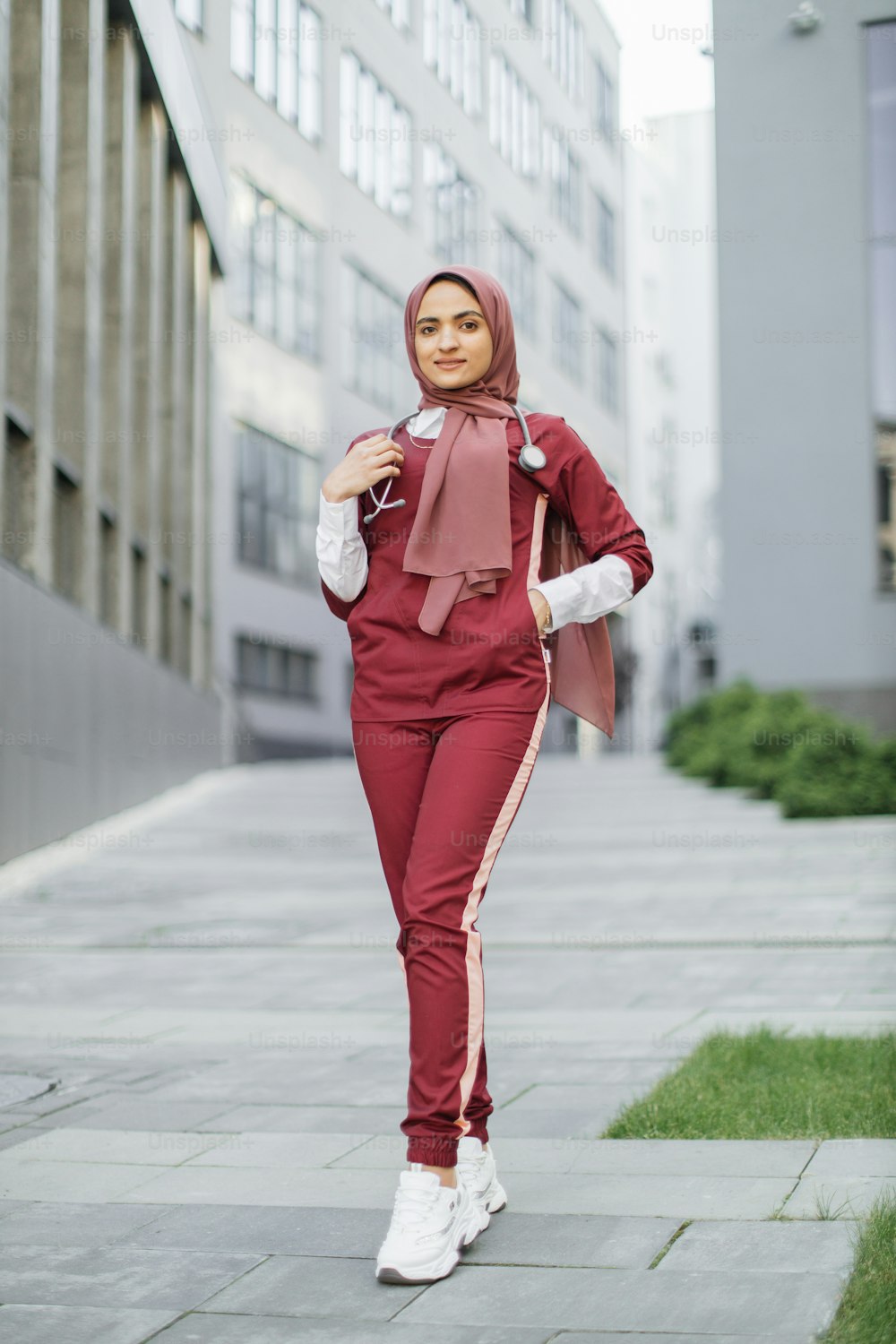 Full length portrait of young Muslim lady doctor, wearing red medical scrubs and treaditional hijab, posing outdoors on the background of modern hospital clinic building