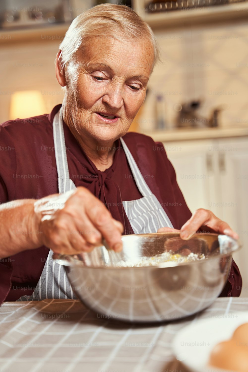 Cooking senior woman holding whisk in right hand and blending dough components inside steel deep bowl