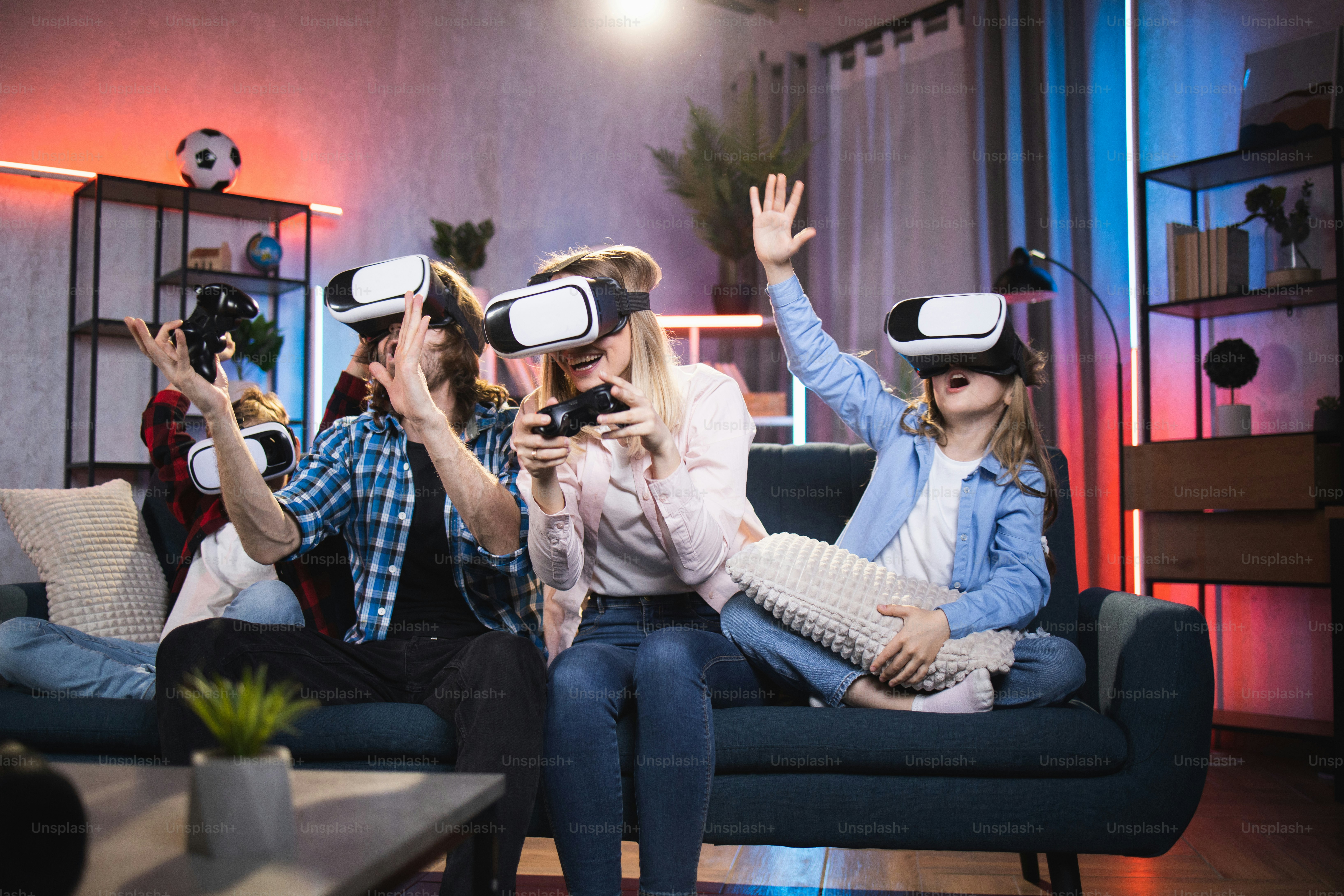 Cheerful kids with their young parents wearing 3D goggles while playing video games at home. Modern family sitting on couch and spending time in virtual reality.