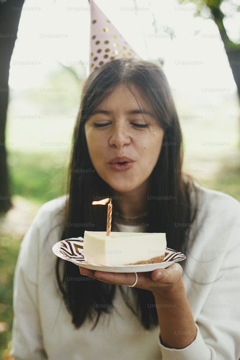 Stylish happy woman in party hat blowing candle on piece of birthday cake and making a wish. Celebrating birthday at picnic party outdoor.