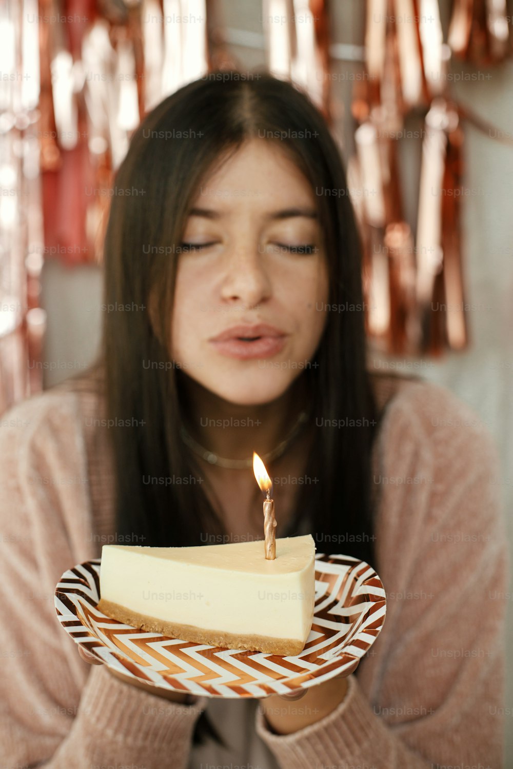 Piece of modern birthday cake with burning candle in hand on background of happy woman and modern rose gold tassel garland in room. Celebrating birthday at home. Make a wish