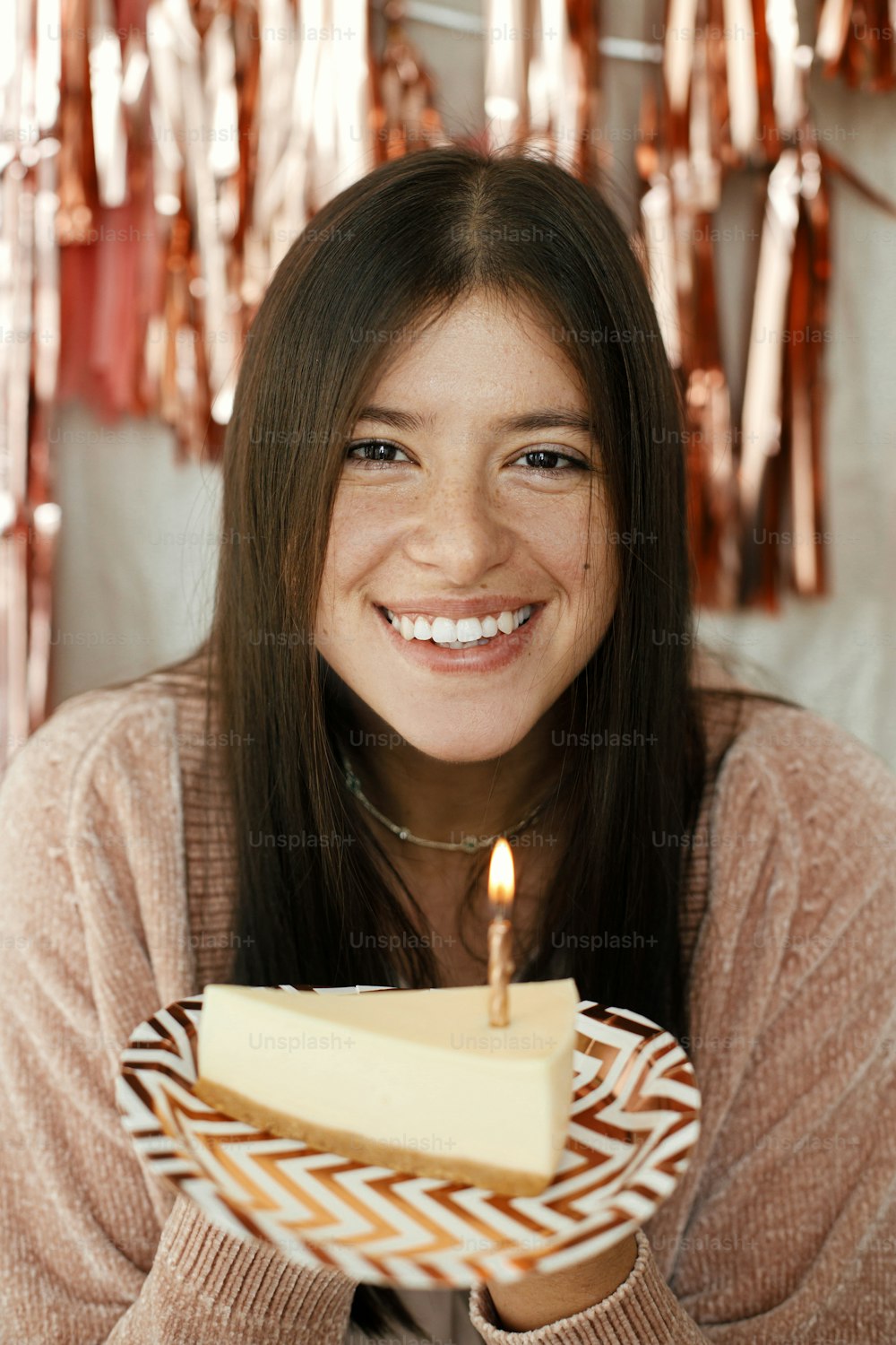 세련된 행복한 여자는 방에 현대 로즈 골드 술 화환을 배경으로 불타는 촛불이 있는 생일 케이크 조각을 들고 있다. 집에서 생일을 축하합니다. 소원을 빌다