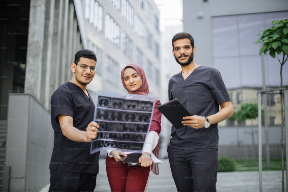 Three doctors, woman in hijab and two men in medical apparel, discussing patient's x-ray tomography scan, walking outside on the background of modern hospital