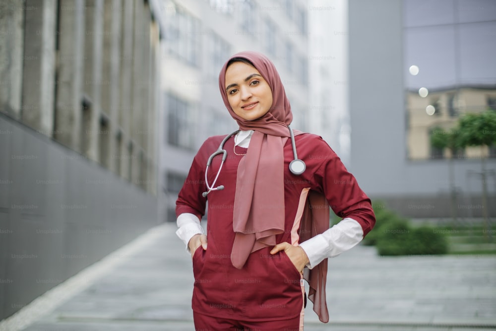 ヒジャーブ、スクラブ、聴診器を身に着けた美しいマレー人女性医師が近代的な診療所の外にいます。屋外に立って、赤いスクラブで、友好的で笑顔の自信に満ちたイスラム教徒の女性医師の接写肖像画
