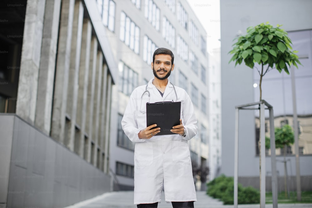 Portrait of young handsome bearded Indian man physician healthcare worker in white coat, standing with clipboard folder in hands outdoors in front of modern hospital or clinic