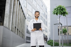 Portrait d’un jeune beau barbu indien médecin soignant en blouse blanche, debout avec un dossier à pince dans les mains à l’extérieur devant un hôpital ou une clinique moderne