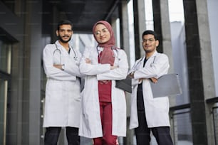 Vista frontal de un equipo altamente calificado de tres jóvenes médicos árabes sauditas seguros de sí mismos, parados afuera del hospital y mirando a la cámara. Bonita dama musulmana con hiyab se para frente a sus dos colegas masculinos