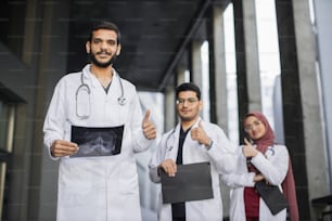 ヘルスケアと人のコンセプト。白い白衣を着たアラブの医療チームが腕を組んで立ち、親指を立てて、ハンサムな男性医師を前に集中させています。