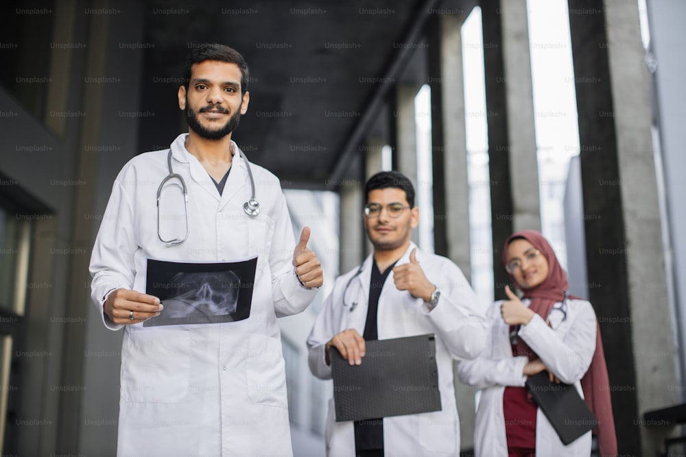 Healthcare and people concept. Happy successful Arab medical team in white lab coats standing with folded arms in a receding row and showing thumbs up, with focus handsome male doctor in front.
