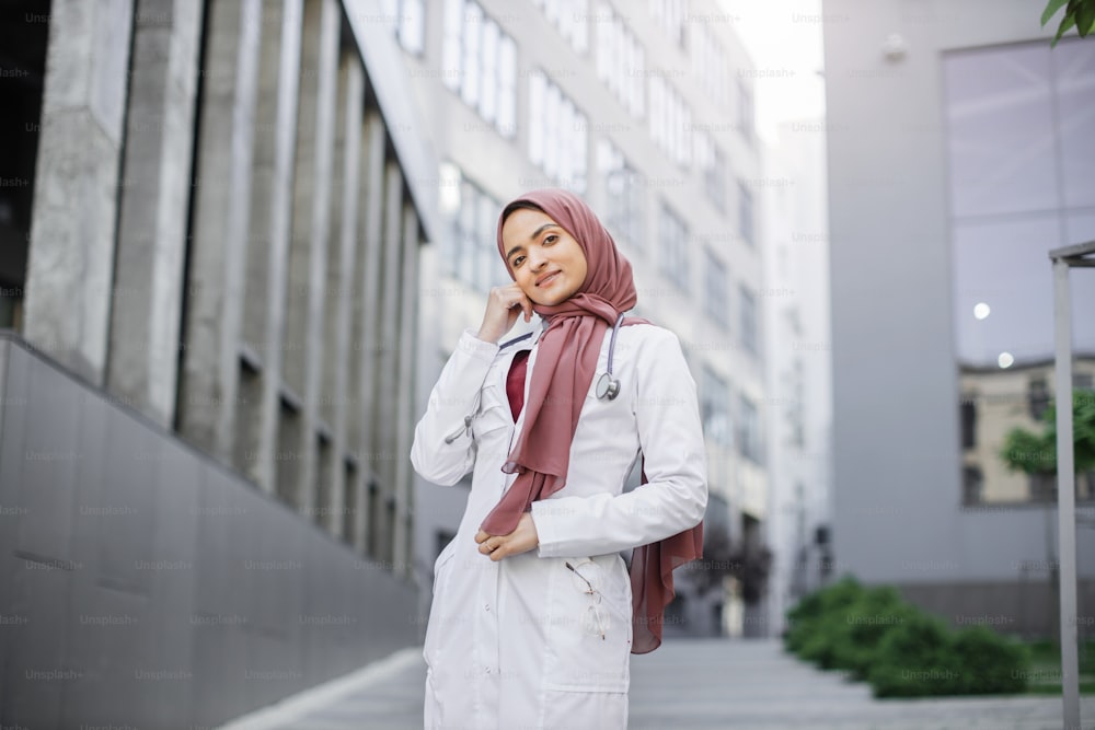 Mitarbeiter im Gesundheitswesen im Nahen Osten. Nahaufnahme des Porträts einer lächelnden, selbstbewussten muslimischen Krankenschwester oder Ärztin, die Hijab trägt und im Freien vor modernen Gebäuden vor der Kamera posiert