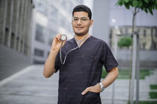 Ritratto di sorridente amichevole maschio arabo indù medico o studente di medicina in piedi fuori dall'ospedale moderno, dimostrando il suo stetoscopio alla macchina fotografica. Messa a fuoco selettiva sul volto dell'uomo