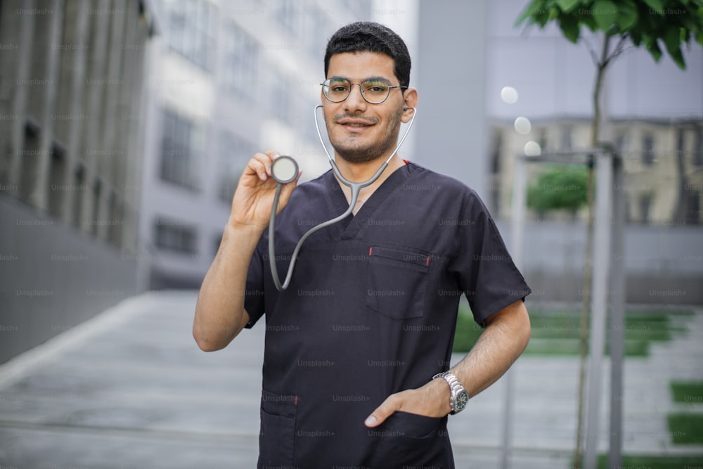 Portrait of smiling friendly male Arab hindu doctor or medical student standing outside modern hospital, demonstrating his stethoscope to camera. Selective focus on man's face