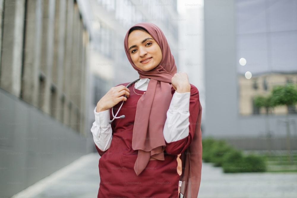 Arab female doctor or medical student, wearing red medical suit and stethoscope, posing and smiling, standing outside on a background of modern university campus or hospital building