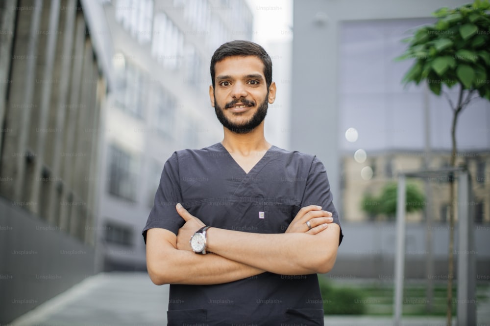 Retrato de cerca de un joven médico árabe profesional masculino, vestido con traje médico gris, sonriendo y mirando a la cámara, de pie al aire libre frente al edificio moderno del hospital