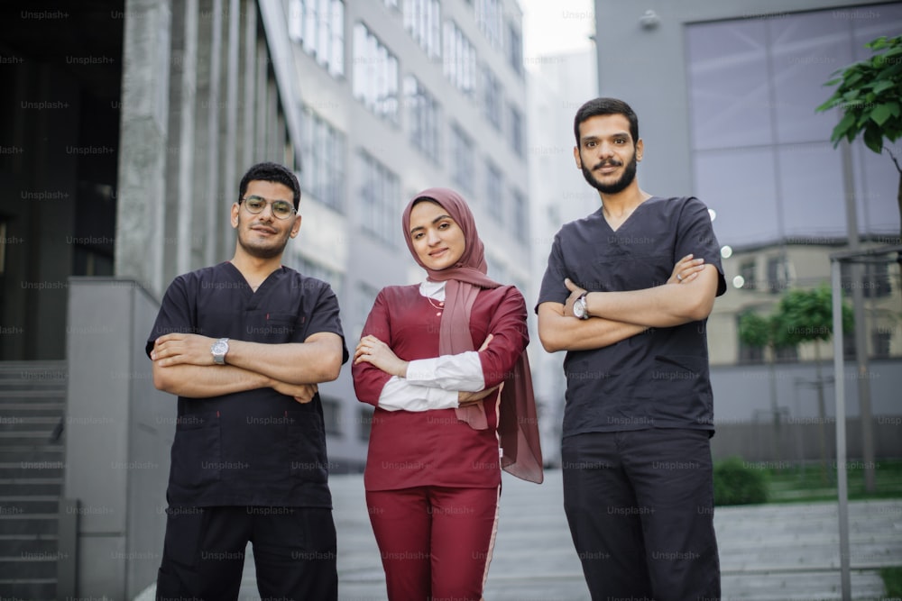 Front view of high-skilled team of three young confident Asian Arab doctors, standing outside the hospital and looking at camera. Pretty Muslim lady in hijab stands between her two male colleagues