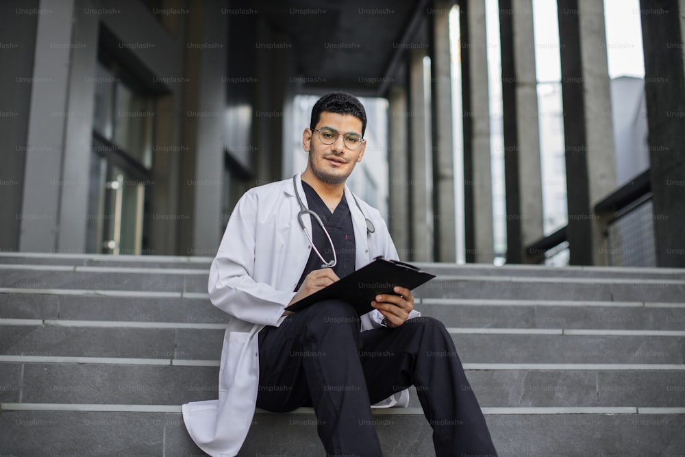 Medical care, insurance, prescription, patient's record concept. Front view of Arabian high-skilled male healthcare professional, sitting on stairs outside hospital and making notes on clipboard