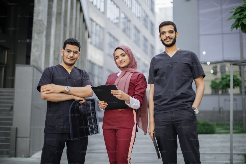 Vista frontal de tres médicos, una mujer musulmana con hiyab y dos hombres árabes, trabajando frente a la clínica. Mujer con portapapeles, hombres con tableta y tomografía computarizada, discutiendo el diagnóstico del paciente