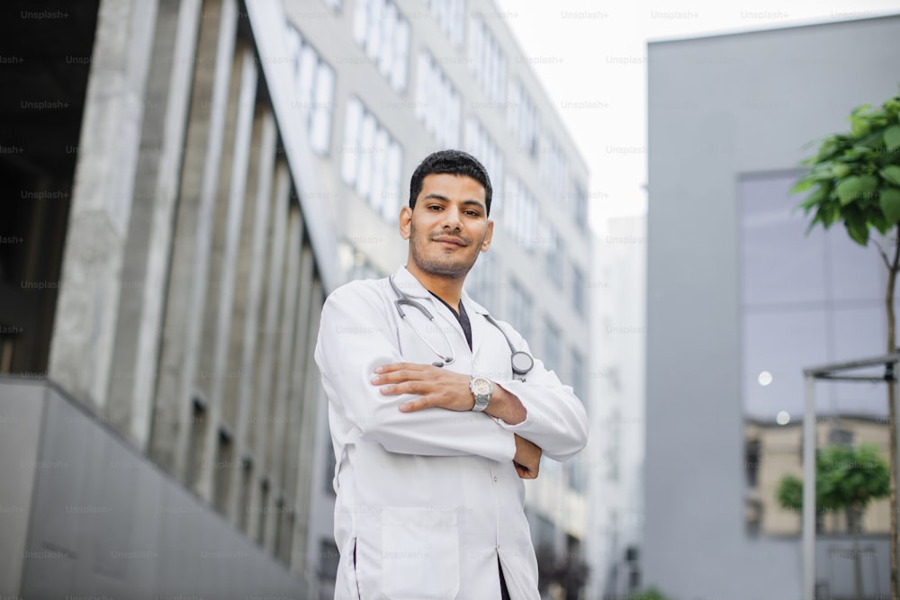 Portrait of young confident Asian Arabian medical doctor standing outside hospital building with arms crossed and stethoscope around neck. Healthcare professional portrait