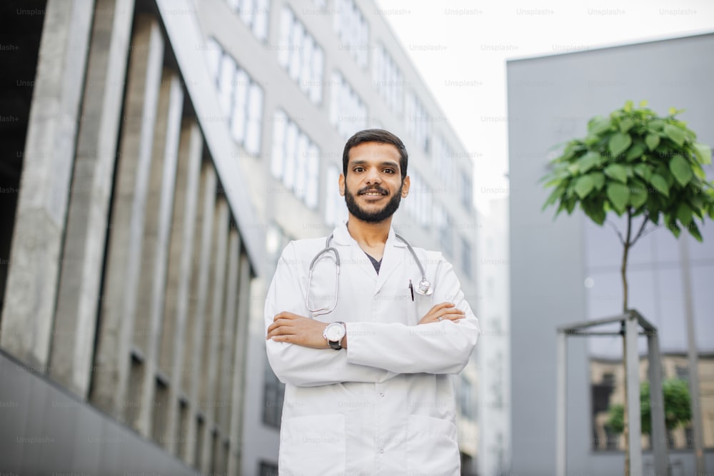 Retrato del joven médico o estudiante árabe asiático masculino en uniforme médico con estetoscopio en el campus universitario moderno de fondo de edificios hospitalarios