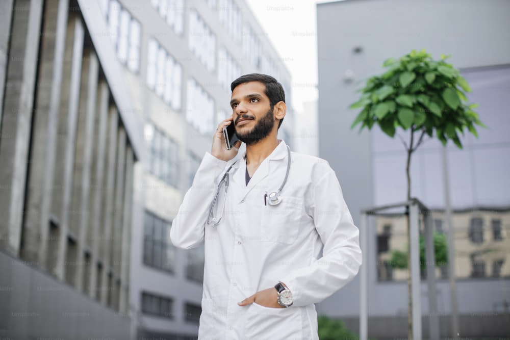 Giovane medico indiano arabo barbuto maschio in piedi fuori dall'ospedale e telefono parlante. Ritratto di piacevole simpatico medico bello che usa il cellulare all'aperto