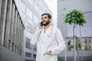 Young male bearded Arabic Indian doctor standing outside hospital and talking phone. Portrait of pleasant likable handsome physician using cellphone outdoors