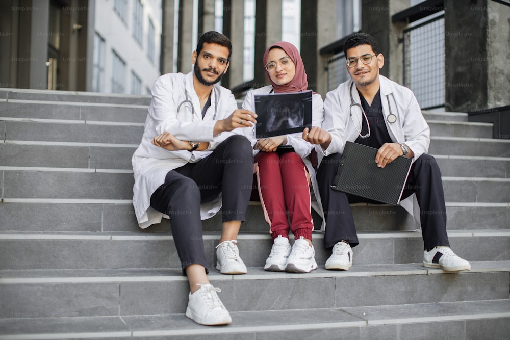 Radiology, ENT and surgery concept. Three young Arabian male and female ENT doctors interns, sitting outdoor on clinic stairs and studying together, brainstorming and discussing skull x-ray