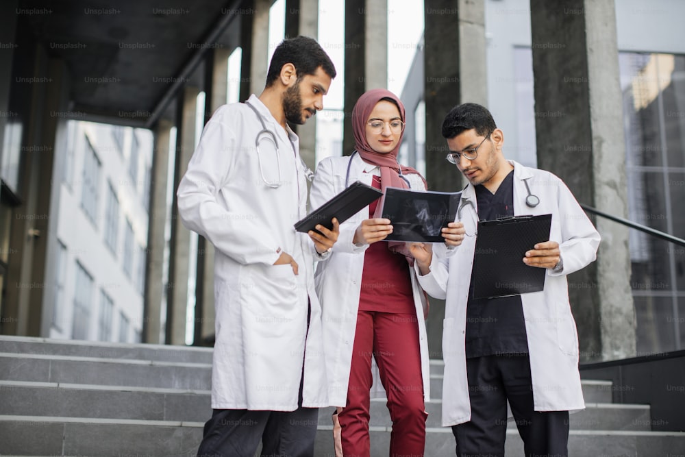 Outdoor shot of team of young skilled confident three Asian Indian doctors, looking at patient's xray, while standing in fonrt of modern stairs of clinic