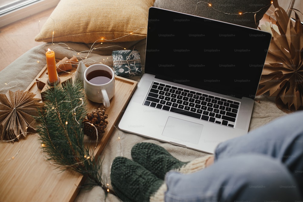 Woman feet in woolen socks on soft bed with laptop, christmas lights, tea, stars, pine, candle and pillows in stylish room. Cozy winter holidays at home. Freelance and blog