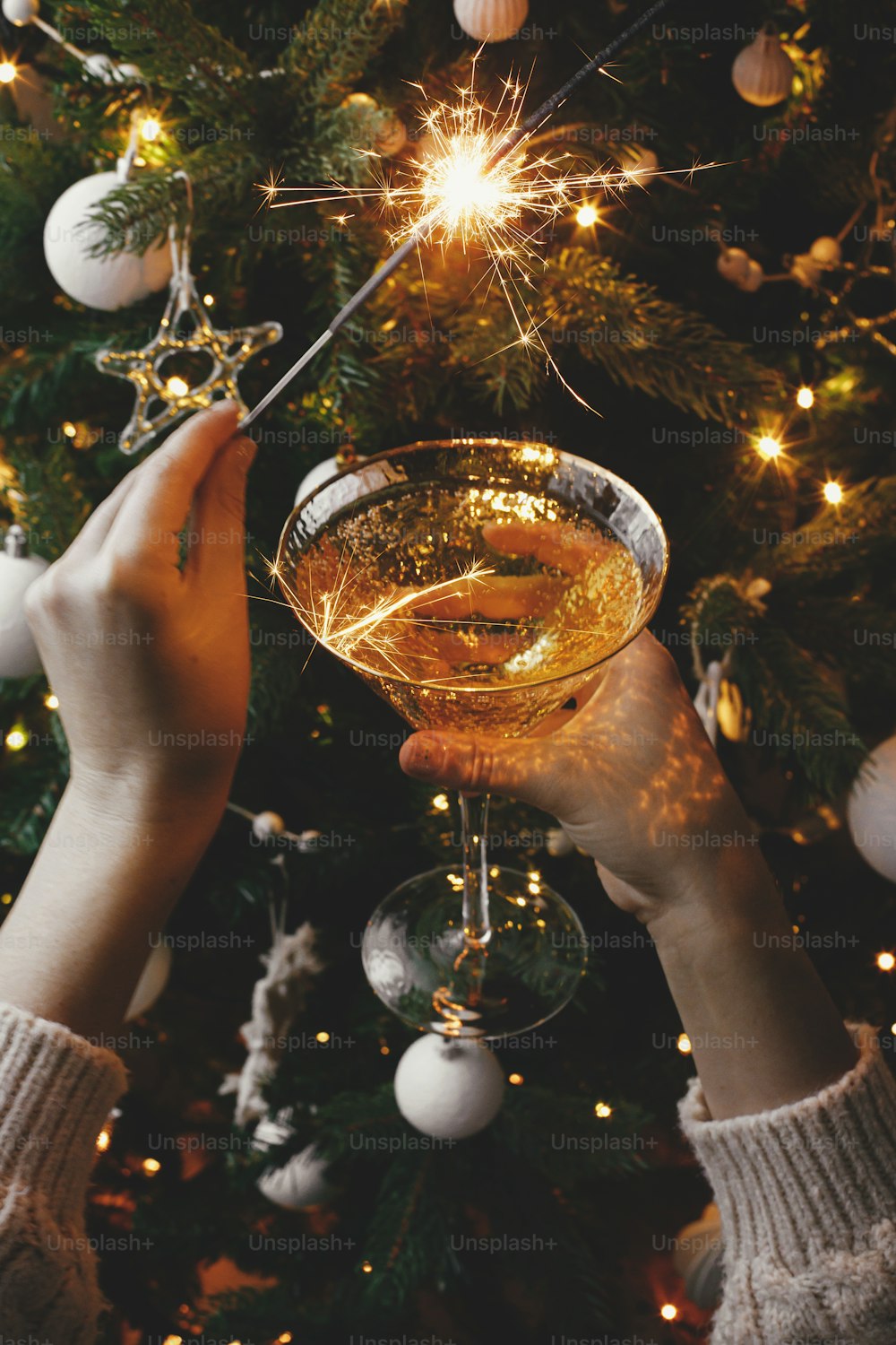 ¡Feliz Año Nuevo! Manos sosteniendo bengala encendida y copa de champán en el fondo de luces del árbol de Navidad. Fuegos artificiales, luz de bengala y bebida en manos de mujer. Momento atmosférico. Fiesta de fin de año