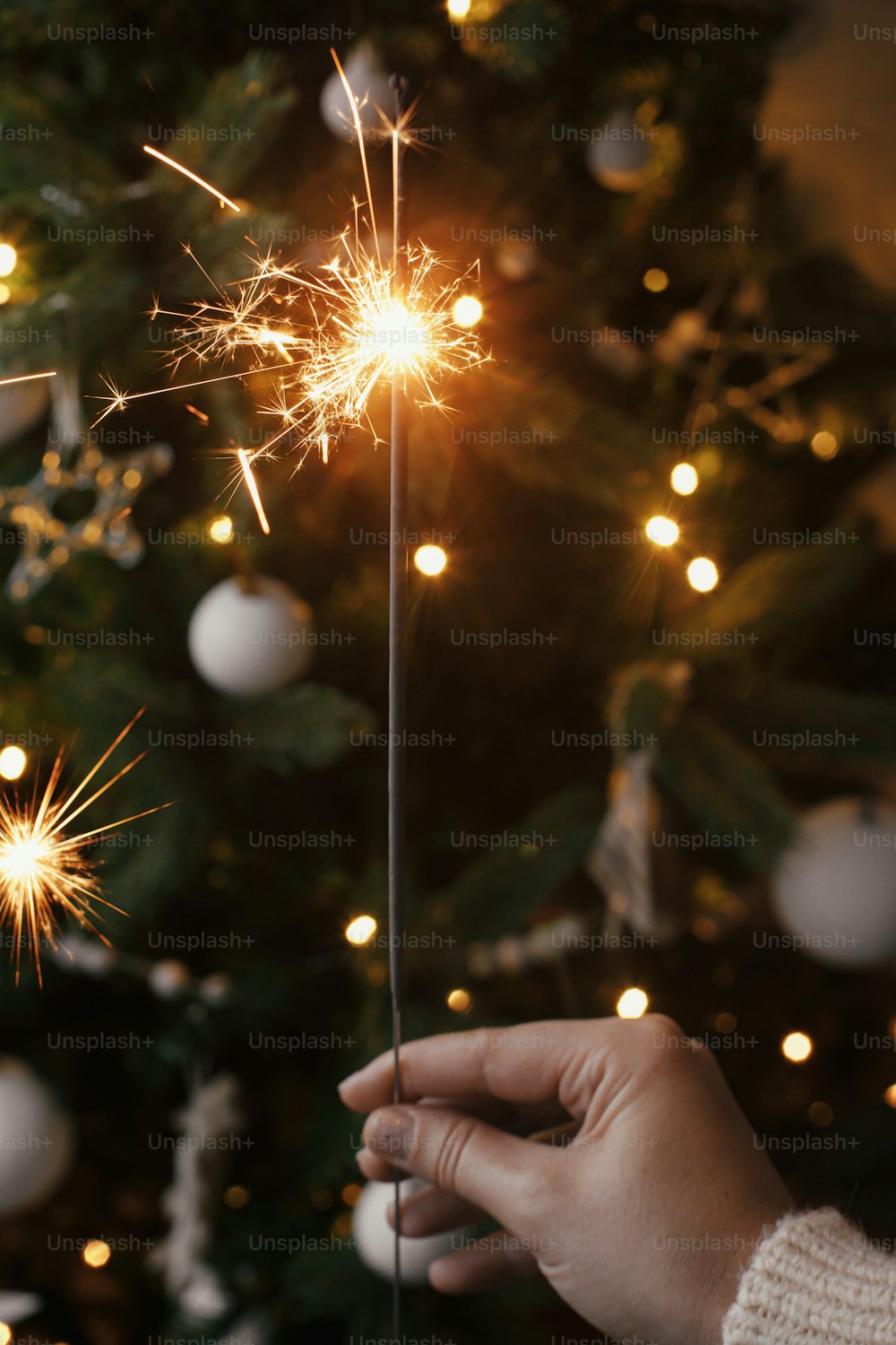 Mano sosteniendo bengala ardiente en el fondo de las luces del árbol de Navidad en la sala de la noche festiva. ¡Feliz Año Nuevo! Fuegos artificiales de bengala que brillan en la mano de la mujer. Espacio para el texto. Momento atmosférico
