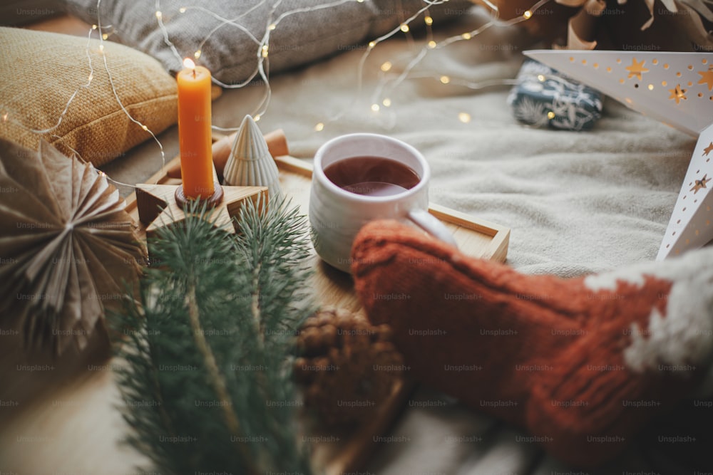Warm cup of tea, woolen socks, christmas stars, golden lights, trees, candle and pillows on soft bed. Cozy moments at scandinavian home. Winter and autumn holidays. Comfort and relax